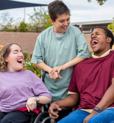 Three people laughing together outdoors, including two wheelchair users.